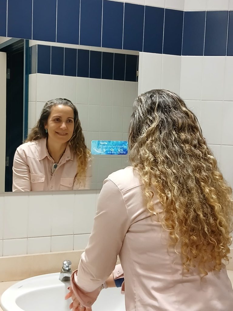 Trabalhadora dos SMAS de Sintra lava as mãos na casa de banho e sorri para o espelho, com autocolante da campanha " Sorri"
